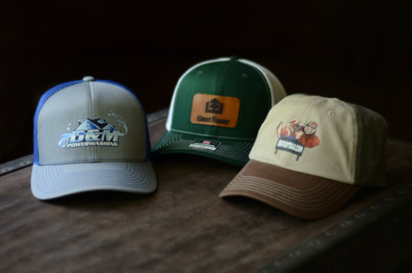 Three baseball caps sit on an old trunk, two with custom business logo prints and one with a leather patch engraved with a business logo.