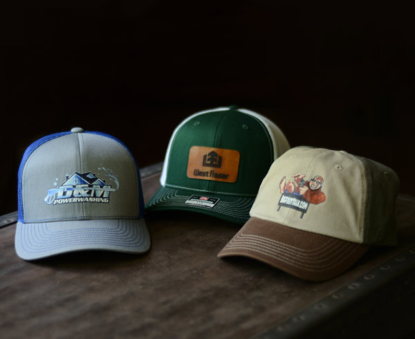 Three baseball caps sit on an old trunk, two with custom business logo prints and one with a leather patch engraved with a business logo.