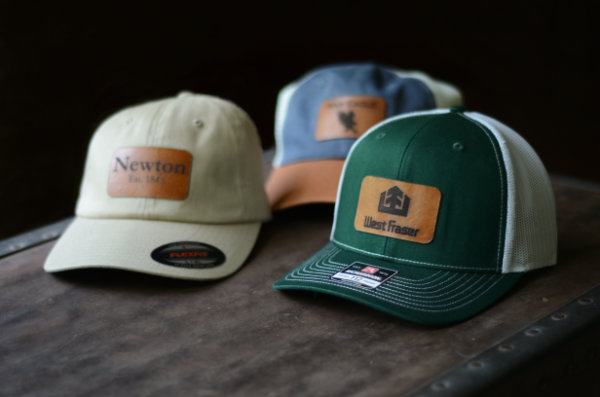 Three baseball caps, each with leather patches engraved with different designs, sit on a vintage trunk.