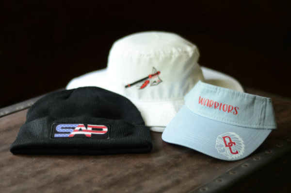 A black beanie, a white boonie hat, and a light blue visor, each decorated with a custom logo, sit on a vintage trunk.